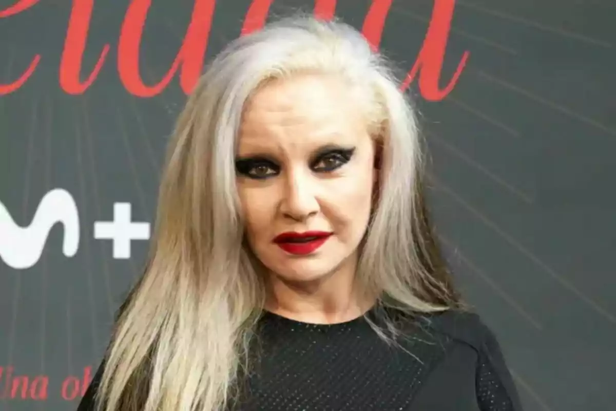 Alaska con cabello rubio y maquillaje oscuro en los ojos y labios rojos frente a un fondo con texto en la presentación de Alaska Revelada. Alaska con cabello rubio y maquillaje oscuro en los ojos y labios rojos frente a un fondo con texto en la presentación de Alaska Revelada.