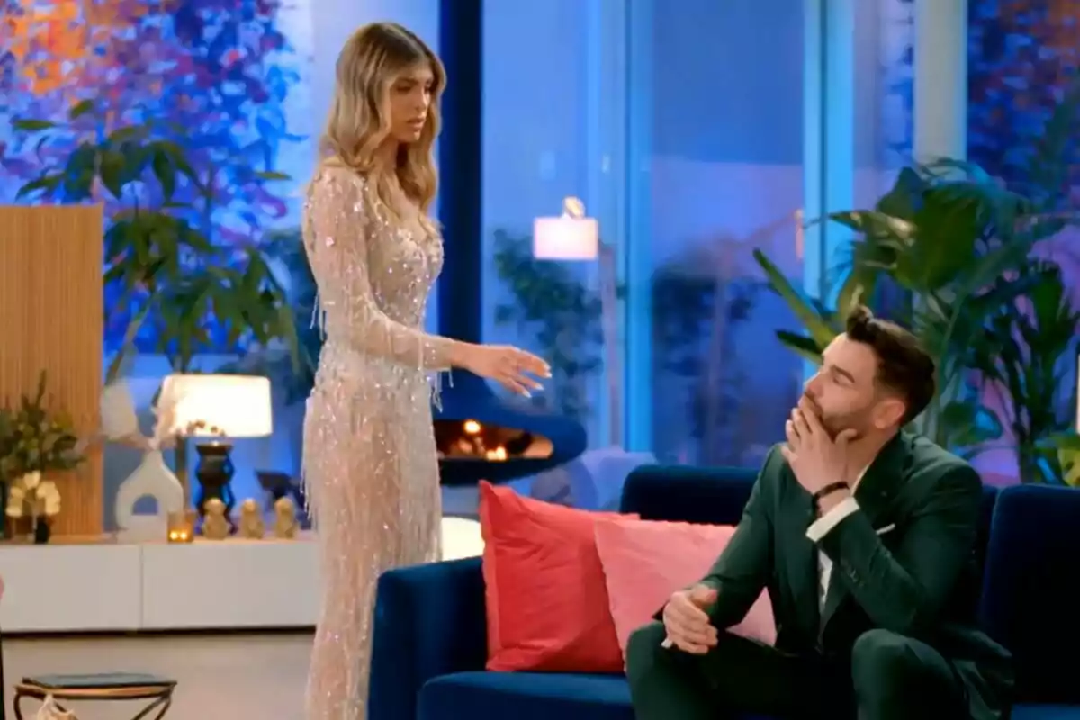 Alba de pie con un vestido brillante parece estar hablando con Gerard sentado en un sofá azul en una sala decorada con plantas y arte colorido en La Isla de las Tentaciones. Alba de pie con un vestido brillante parece estar hablando con Gerard sentado en un sofá azul en una sala decorada con plantas y arte colorido en La Isla de las Tentaciones.