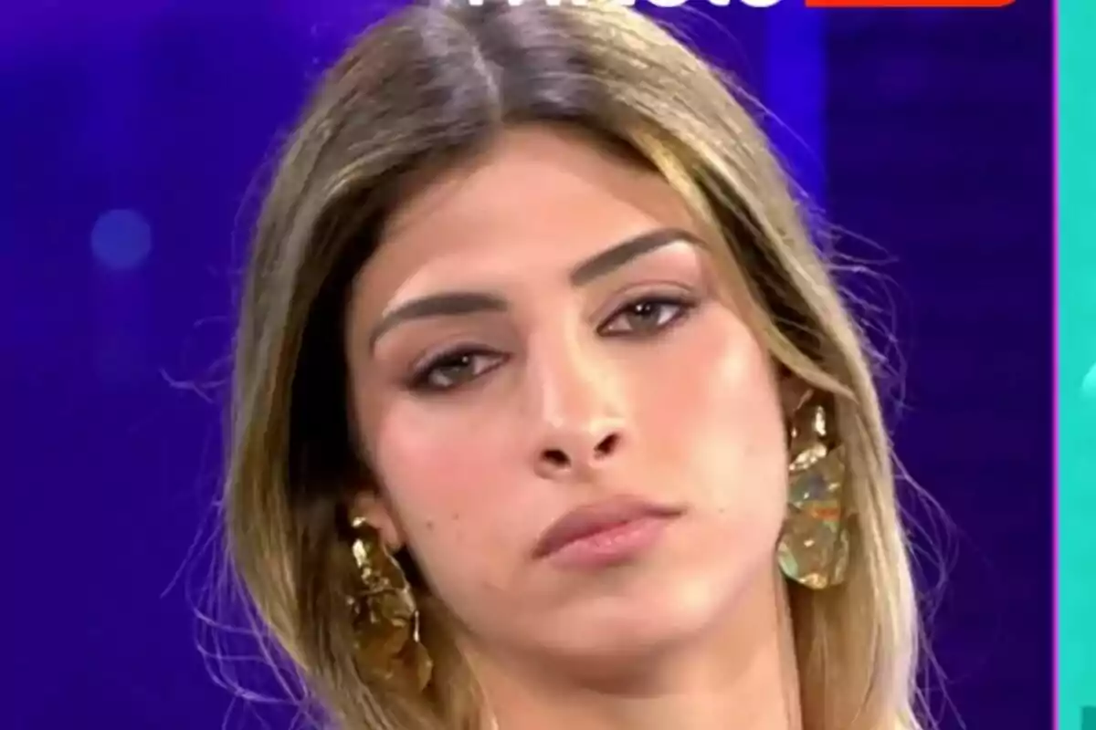 Alba Rodríguez con cabello largo y aretes grandes mira hacia adelante con una expresión seria en un fondo azul en Los vecinos de la casa de al lado.