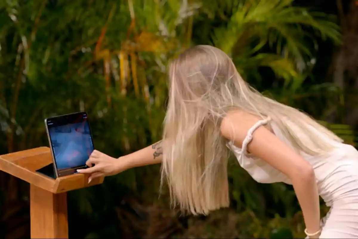 Alba con cabello largo y rubio interactúa con una tablet en un soporte de madera, rodeada de vegetación en La Isla de las Tentaciones. Alba con cabello largo y rubio interactúa con una tablet en un soporte de madera, rodeada de vegetación en La Isla de las Tentaciones.