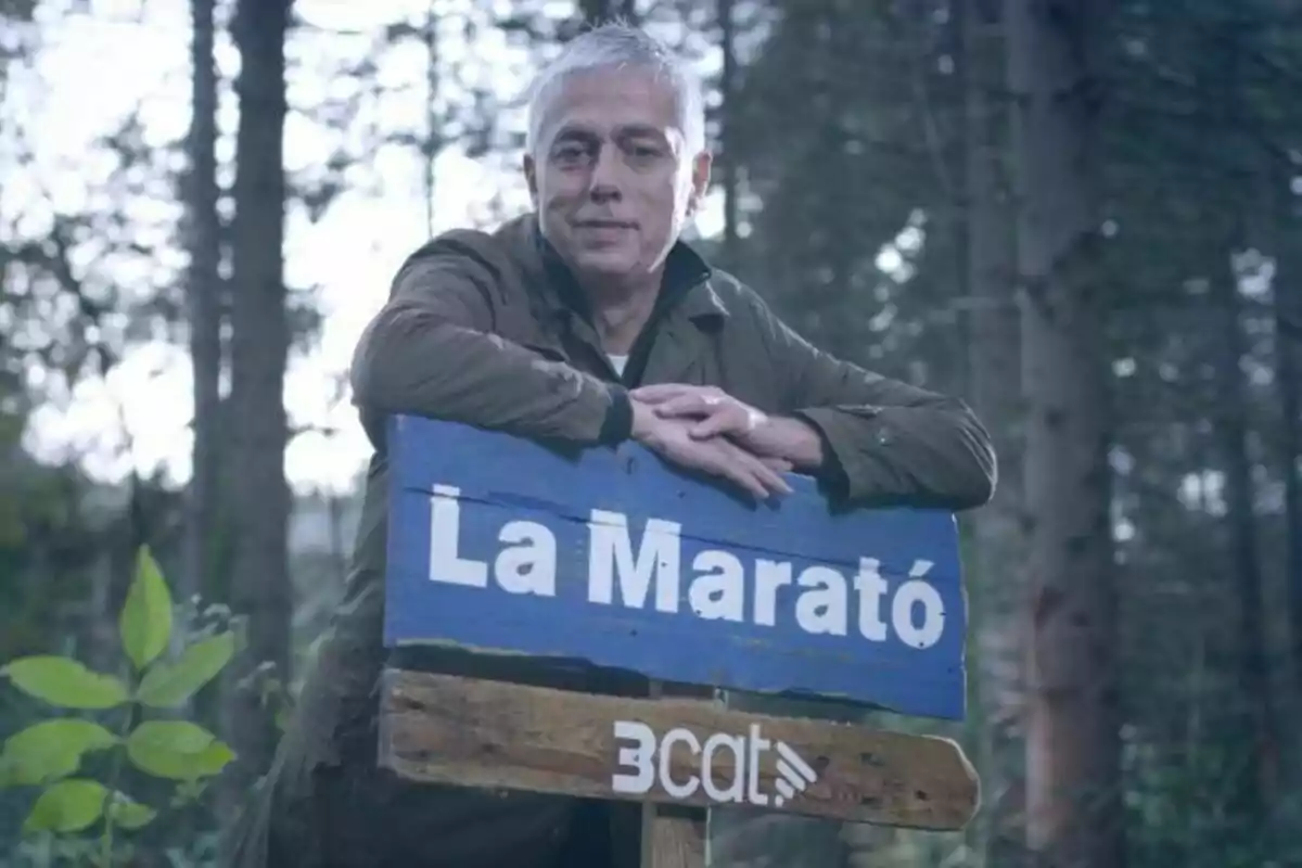 Albert Om de cabello canoso apoya sus brazos sobre un cartel azul que dice "La Marató" en un entorno boscoso.
