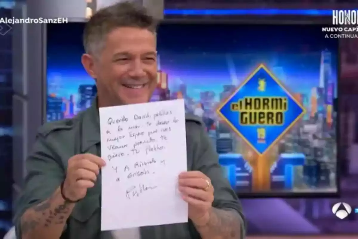 Alejandro Sanz somrient sosté un full de paper amb un missatge escrit a mà, mentre és en un plató de televisió amb el logotip d' Alejandro Sanz somrient sosté un full de paper amb un missatge escrit a mà, mentre és en un plató de televisió amb el logotip d'