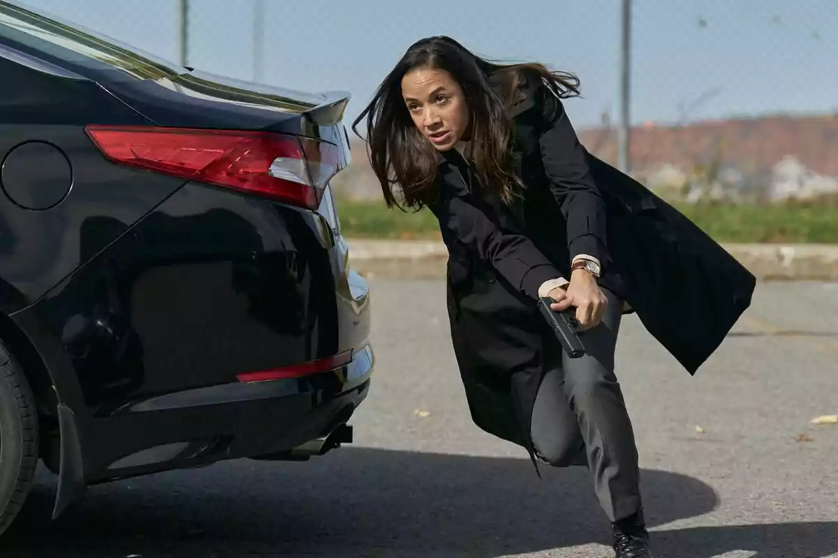 Captura de Dania Ramirez agachada junto a un coche negro sosteniendo una pistola en una escena al aire libre en Alert: Unidad de Personas Desaparecidas Captura de Dania Ramirez agachada junto a un coche negro sosteniendo una pistola en una escena al aire libre en Alert: Unidad de Personas Desaparecidas