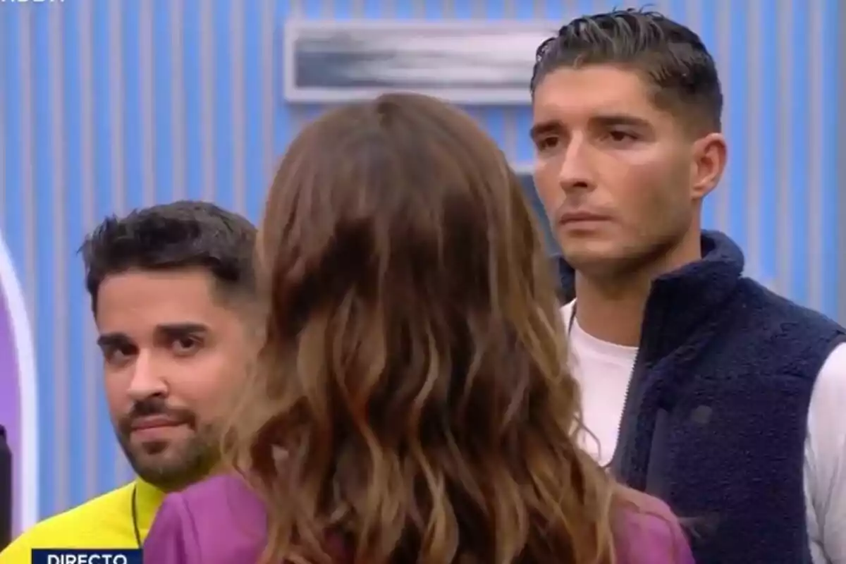 Tres persones a la casa de GH DÚO amb fons de ratlles blaves, Miguel Frigenti i Álex Ghita miren endavant mentre Elena Rodríguez està d'esquena.