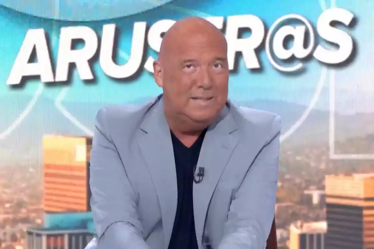 Alfonso Arús calb amb jaqueta grisa apareix davant d'un fons amb el text 