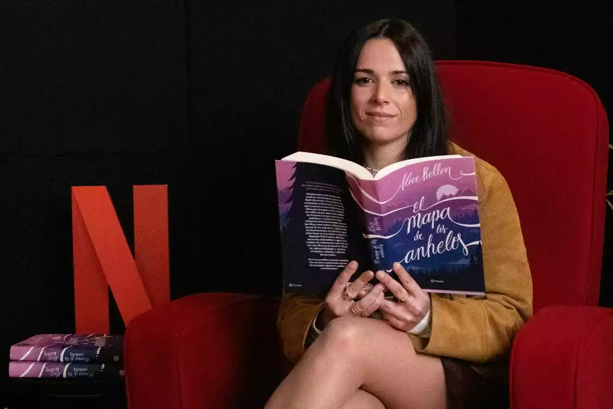 Alice Kellen sentada en un sillón rojo sosteniendo un libro titulado "El mapa de los anhelos" con un fondo negro y una letra "N" roja de Netflix al lado.