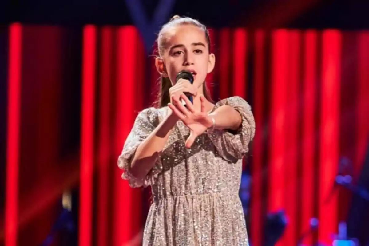 Fotografía de Alira Moya cantando en un escenario de La Voz Kids con cortinas rojas de fondo. Fotografía de Alira Moya cantando en un escenario de La Voz Kids con cortinas rojas de fondo.