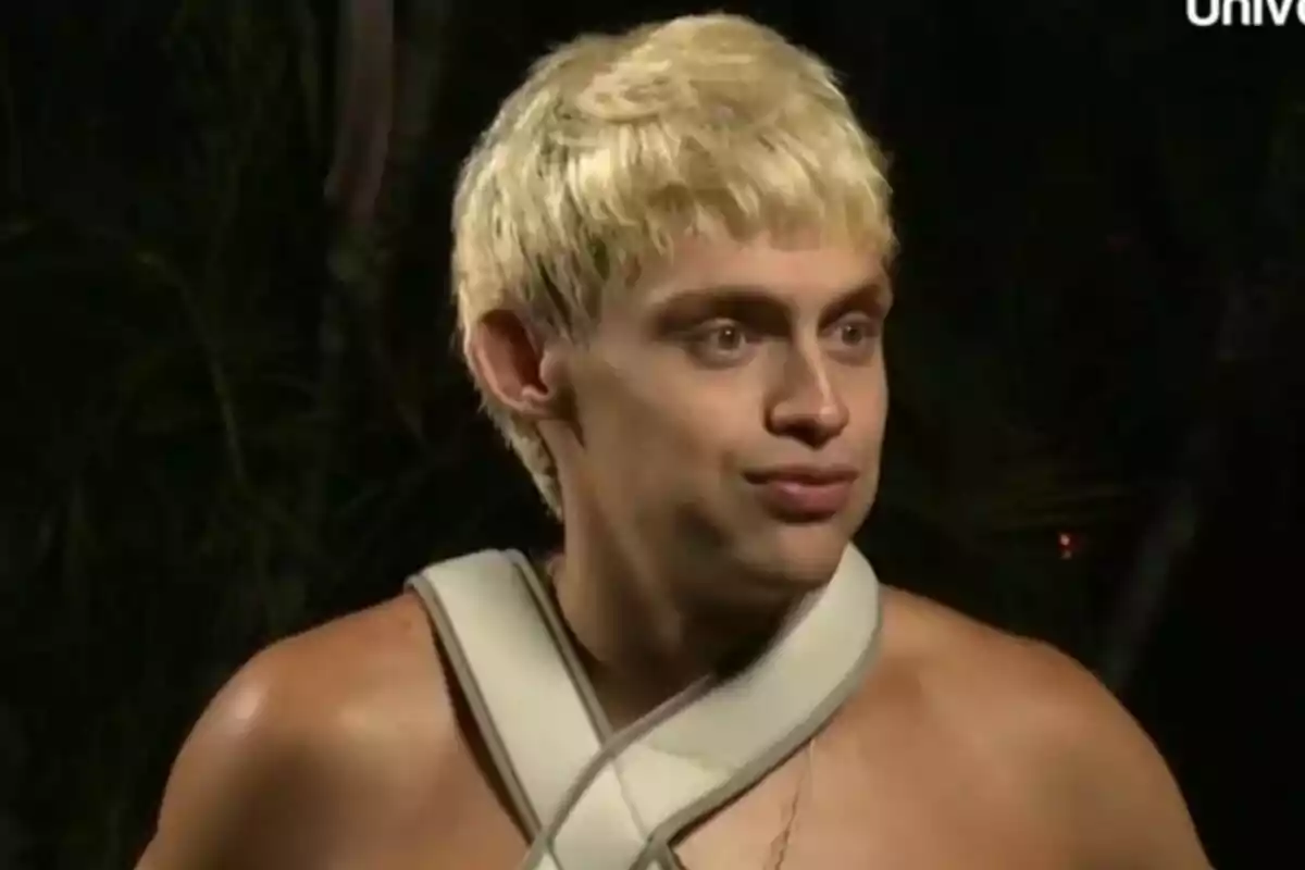 Almácor con cabello rubio y expresión sorprendida lleva un vendaje alrededor del cuello en un entorno oscuro en Supervivientes.