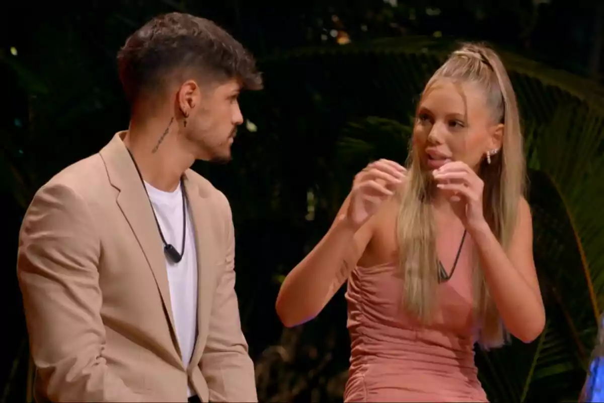 Álvaro y Alba sentadas conversando, una mujer con vestido rosa gesticulando y un hombre con chaqueta beige mirándola, en un entorno con vegetación en La Isla de las Tentaciones. Álvaro y Alba sentadas conversando, una mujer con vestido rosa gesticulando y un hombre con chaqueta beige mirándola, en un entorno con vegetación en La Isla de las Tentaciones.
