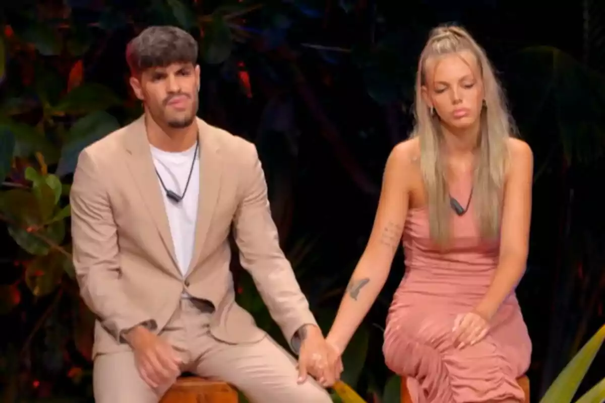 Álvaro y Alba sentada en un entorno tropical, ambos con expresiones serias, sosteniéndose de la mano; él lleva un traje beige y ella un vestido rosa en La Isla de las Tentaciones. Álvaro y Alba sentada en un entorno tropical, ambos con expresiones serias, sosteniéndose de la mano; él lleva un traje beige y ella un vestido rosa en La Isla de las Tentaciones.