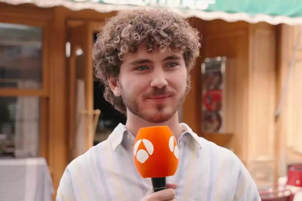 Álvaro de Juana con cabello rizado sostiene un micrófono naranja frente a un edificio de madera en Amar es para siempre. Álvaro de Juana con cabello rizado sostiene un micrófono naranja frente a un edificio de madera en Amar es para siempre.
