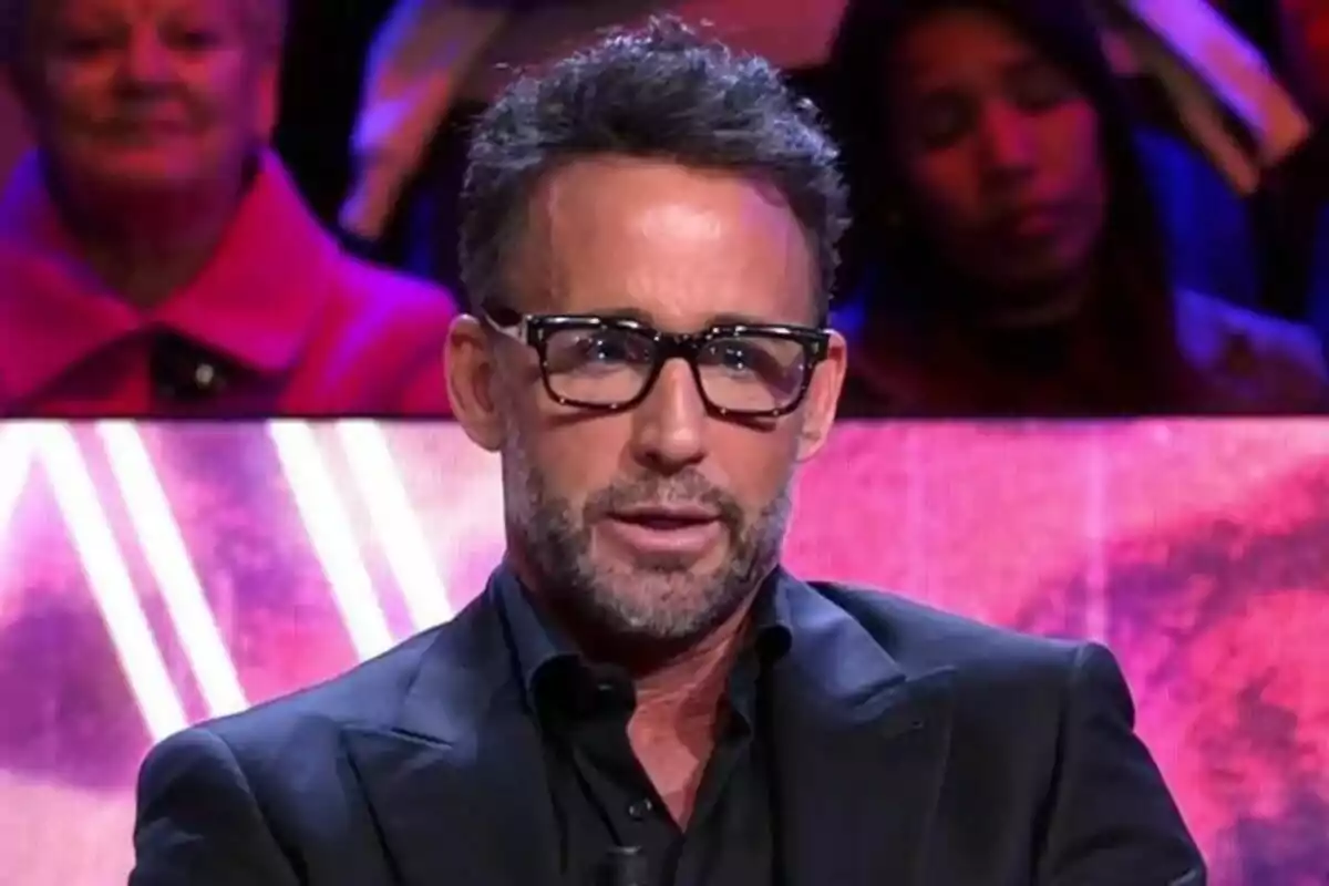 Álvaro Muñoz Escassi con gafas y barba está sentado en De Viernes, con un fondo colorido y personas desenfocadas detrás. Álvaro Muñoz Escassi con gafas y barba está sentado en De Viernes, con un fondo colorido y personas desenfocadas detrás.