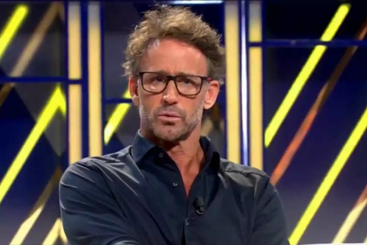 Captura de Álvaro Muñoz Escassi en el plató de De Viernes, con gafas y camisa oscura en un set de televisión con luces de neón amarillas y azules. Captura de Álvaro Muñoz Escassi en el plató de De Viernes, con gafas y camisa oscura en un set de televisión con luces de neón amarillas y azules.
