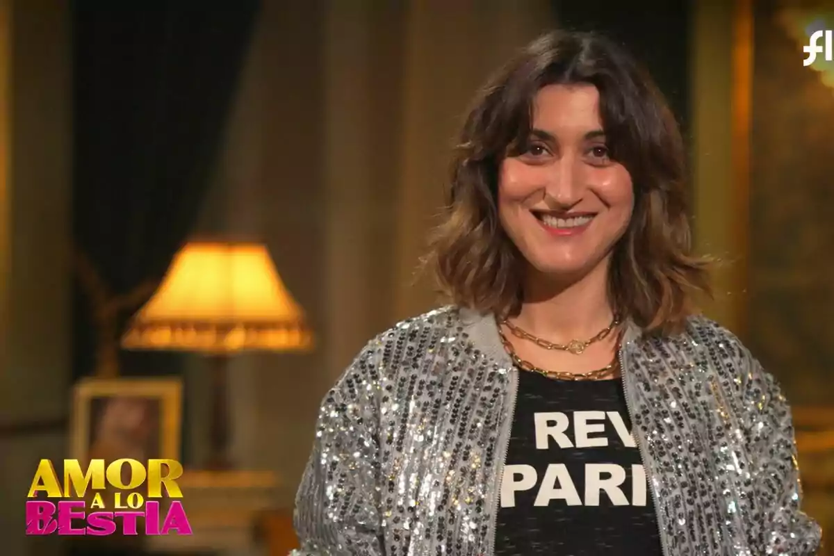 Susi Caramelo sonriente con una chaqueta brillante y una camiseta negra, en un ambiente interior con una lámpara de fondo y el logo de Susi Caramelo sonriente con una chaqueta brillante y una camiseta negra, en un ambiente interior con una lámpara de fondo y el logo de