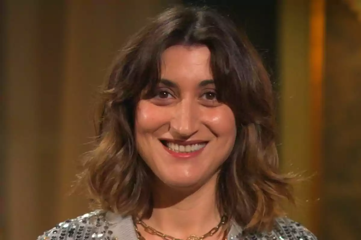 Susi Caramelo sonriendo con cabello castaño y ondulado, usando un collar dorado y una chaqueta con lentejuelas en Amor a lo bestia.