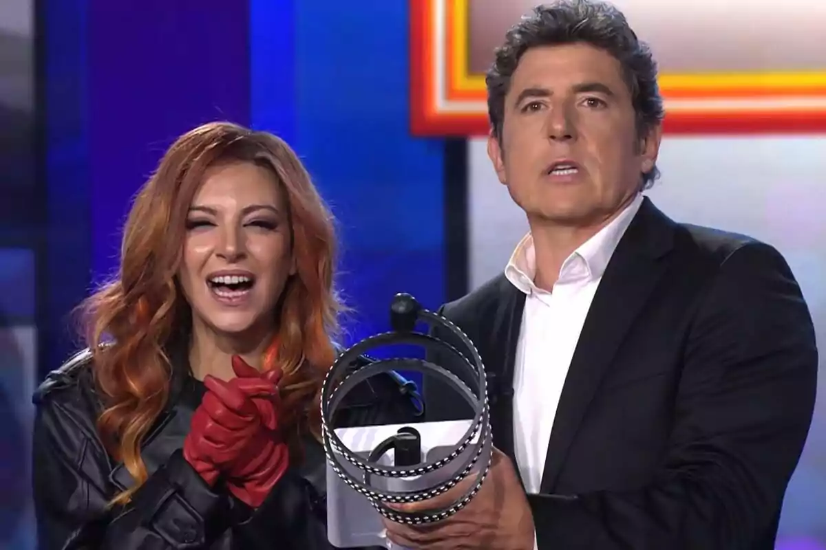 Dos personas en un set de televisión de Tu Cara Me Suena, Ana Guerra sonriente con guantes rojos y Manel Fuentes de traje sosteniendo un objeto metálico circular
