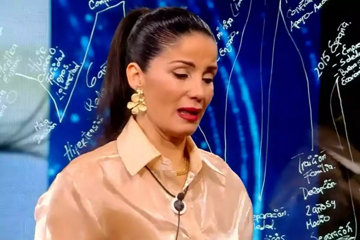 Ana Herminia amb cabell fosc recollit i pendents daurats grans està parlant a GH DÚO amb un fons blau que té text escrit en blanc.