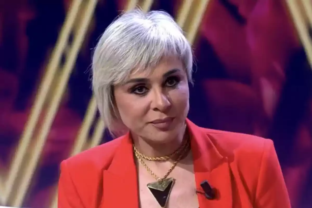 Ana María Aldón con cabello corto y rubio vestida con un traje rojo y un collar dorado en un ambiente iluminado.