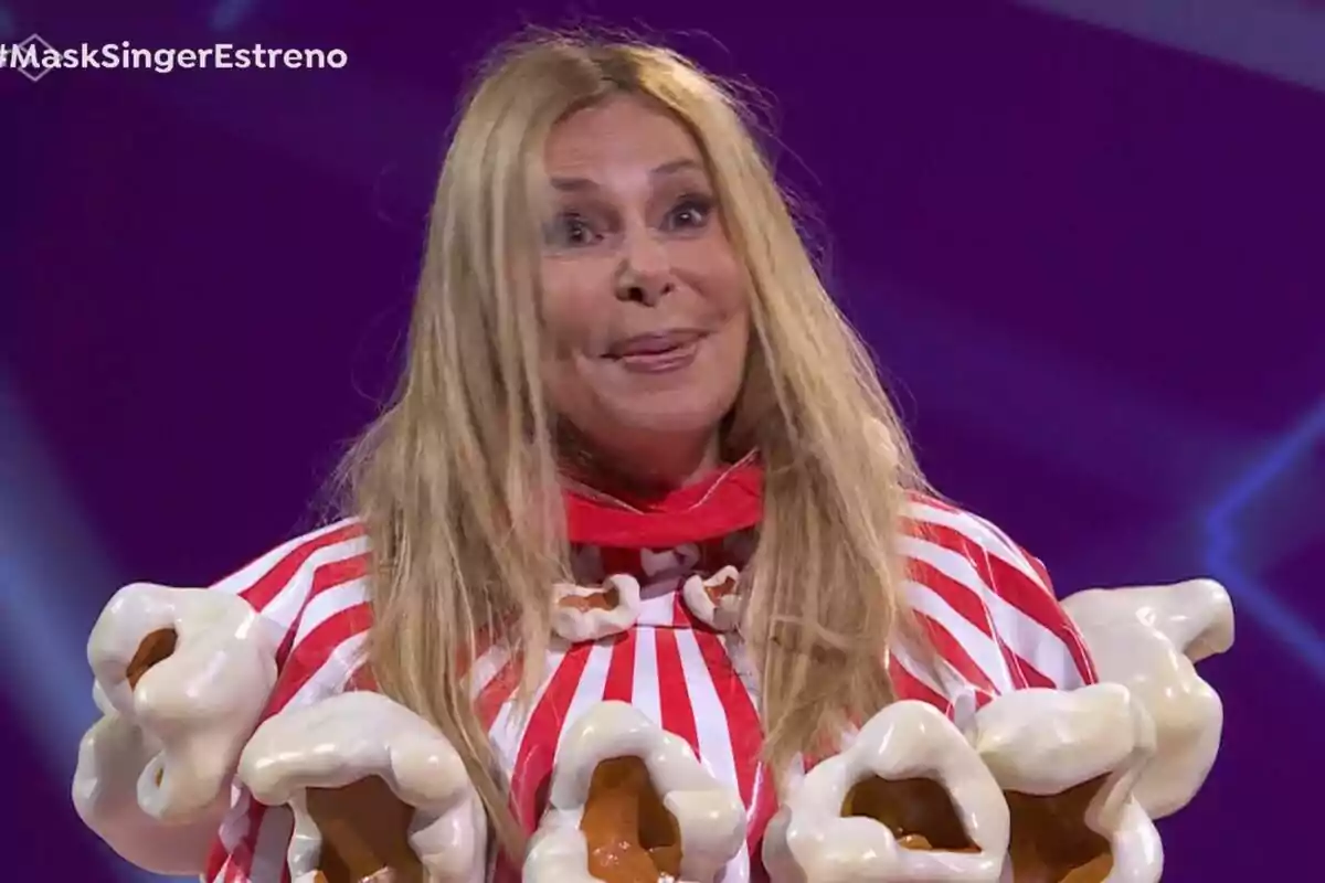 Ana Obregón con cabello rubio y largo está vestida con un disfraz colorido que parece tener elementos de palomitas de maíz, en un escenario con fondo morado en Mask Singer. Ana Obregón con cabello rubio y largo está vestida con un disfraz colorido que parece tener elementos de palomitas de maíz, en un escenario con fondo morado en Mask Singer.