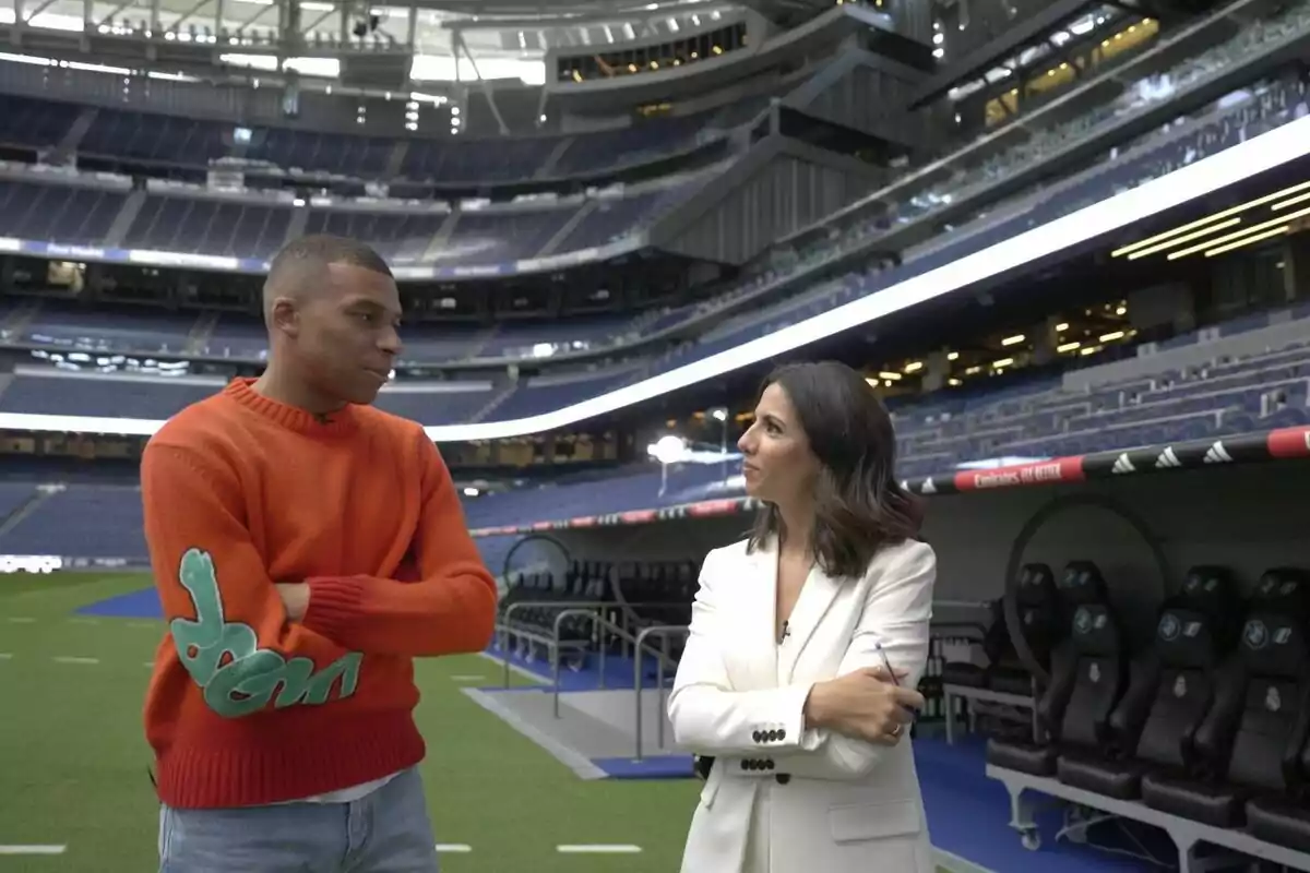 Ana Pastor y Kylian Mbappé conversan en un estadio de fútbol vacío, una lleva un suéter naranja y la otra un traje blanco en El Objetivo.
