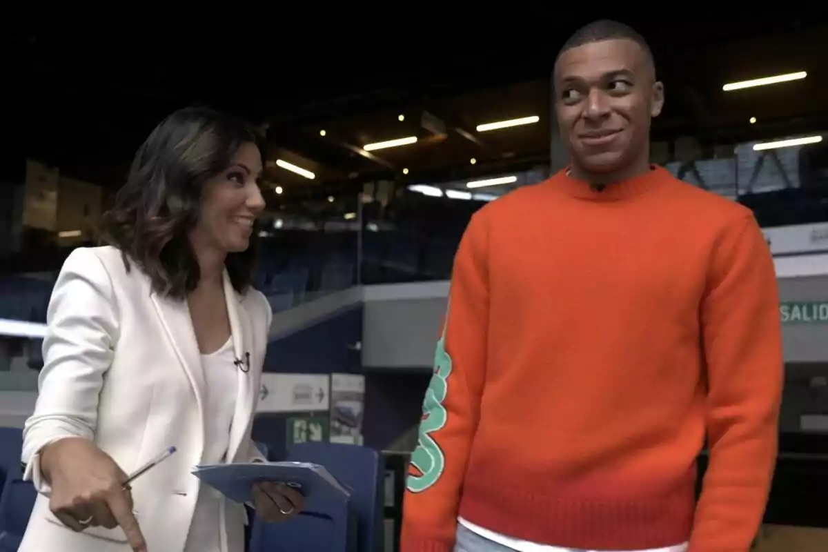 Ana Pastor somrient en vestit blanc sosté papers i un bolígraf mentre conversa amb Kylian Mbappé que porta un jersei taronja en un entorn interior a El Objetivo.