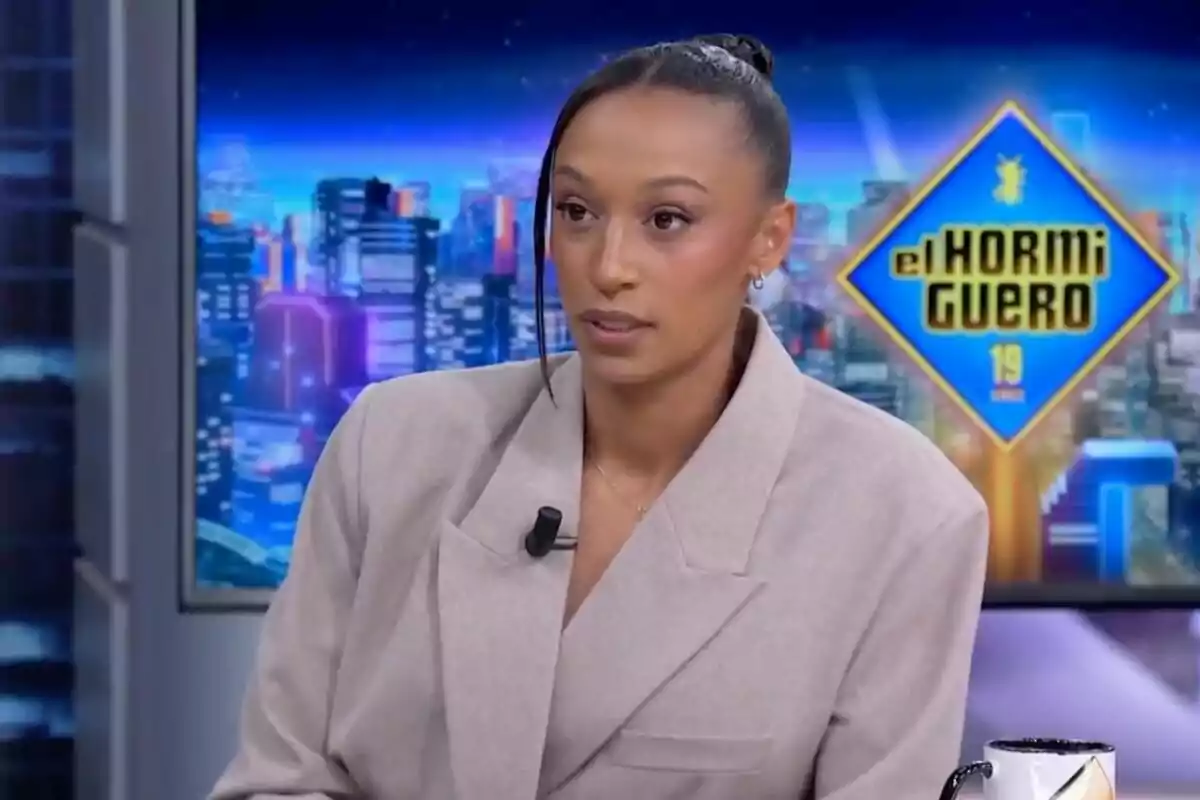 Ana Peleteiro con traje claro está sentada en un set de televisión con un fondo de ciudad iluminada y un cartel que dice Ana Peleteiro con traje claro está sentada en un set de televisión con un fondo de ciudad iluminada y un cartel que dice