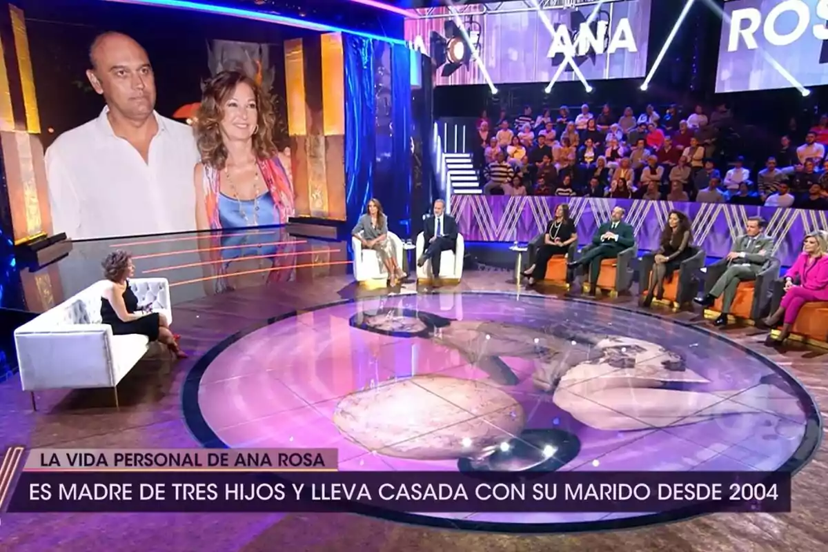 Un plató de De Viernes con varias personas sentadas en un círculo, una gran pantalla muestra a una pareja y hay un texto que dice Un plató de De Viernes con varias personas sentadas en un círculo, una gran pantalla muestra a una pareja y hay un texto que dice