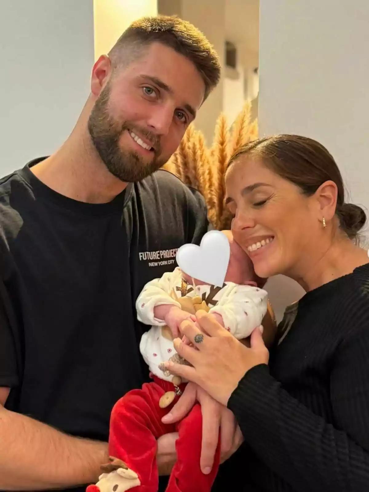 Una pareja sonriente sostiene a un bebé con un corazón blanco cubriendo su rostro. Una pareja sonriente sostiene a un bebé con un corazón blanco cubriendo su rostro.