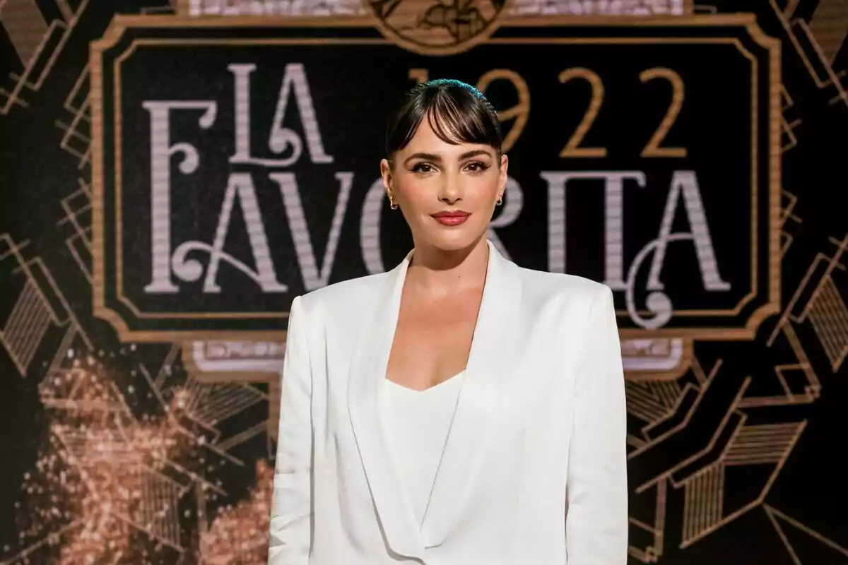 Andrea Duro amb vestit blanc posant davant d'un fons decoratiu que diu 