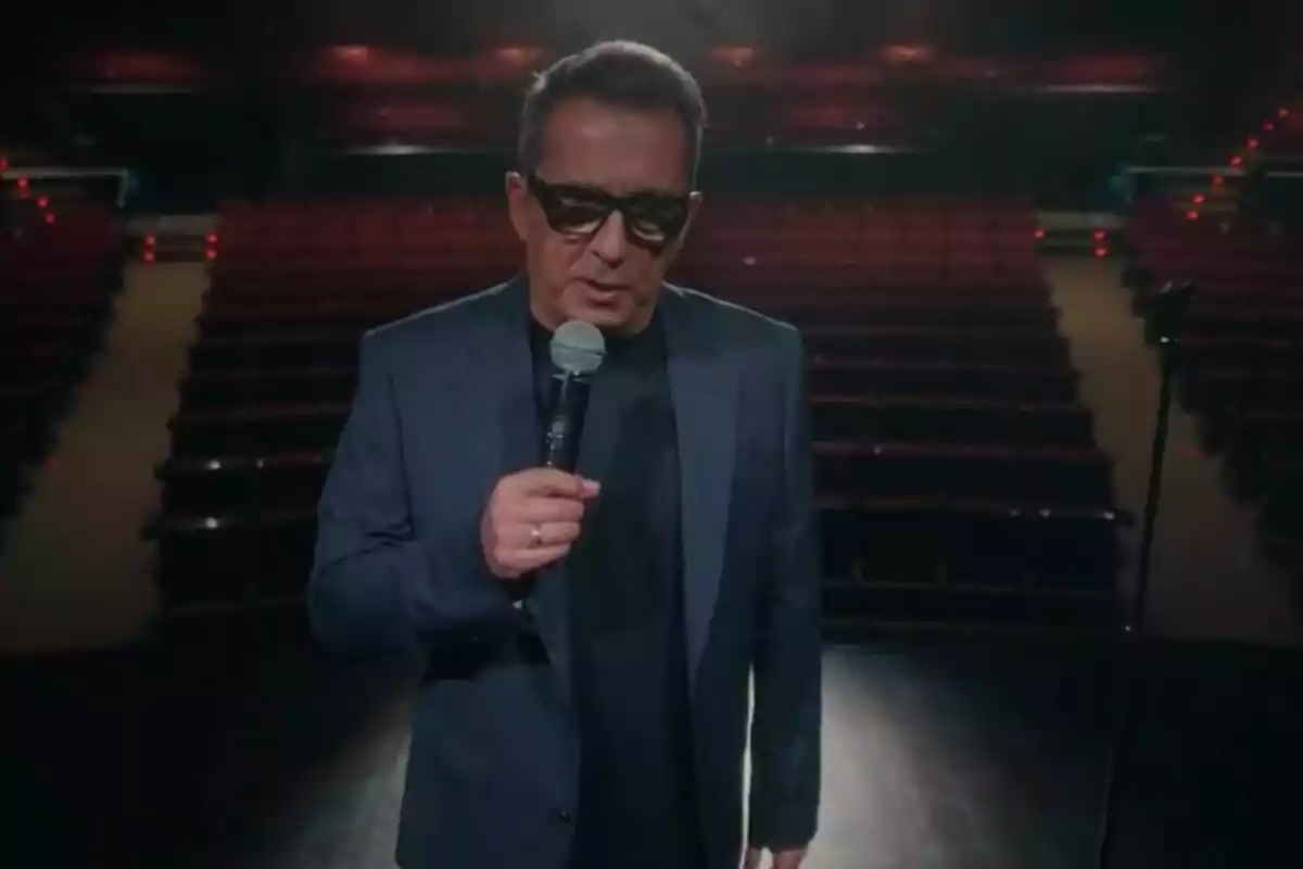 Andreu Buenafuente con gafas de sol y traje oscuro sostiene un micrófono en un escenario de teatro vacío en Futuro Imperfecto. Andreu Buenafuente con gafas de sol y traje oscuro sostiene un micrófono en un escenario de teatro vacío en Futuro Imperfecto.
