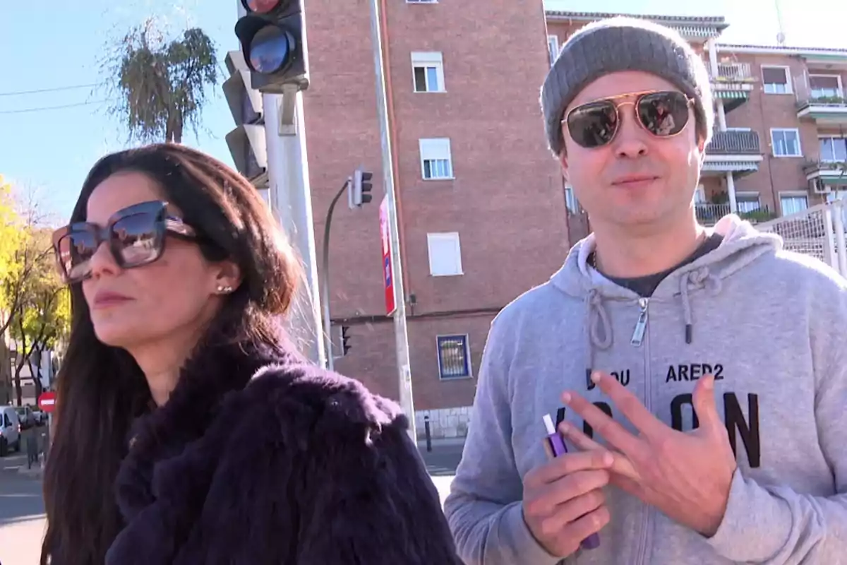 Ángel Cristo y Ana Herminia con gafas de sol en una calle urbana junto a un semáforo y edificios de fondo. Ángel Cristo y Ana Herminia con gafas de sol en una calle urbana junto a un semáforo y edificios de fondo.