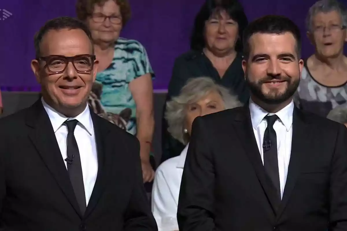 Àngel Llàcer i Ricard Ustrell vestits amb vestit i corbata negra somriuen davant d'una audiència en un estudi de televisió de Col·lapse.