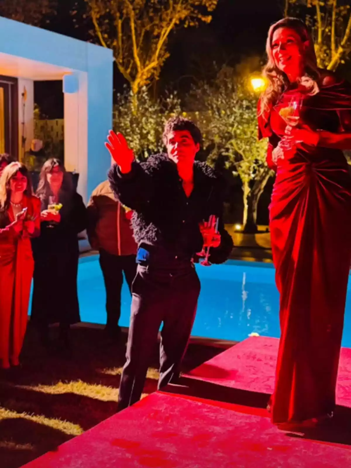 Anne Igartiburu con un vestido rojo y una copa en la mano está de pie sobre una plataforma alfombrada, mientras Javi Ambrossi con chaqueta oscura y también con una copa parece estar hablando o señalando algo, con un grupo de personas al fondo cerca de una piscina iluminada. Anne Igartiburu con un vestido rojo y una copa en la mano está de pie sobre una plataforma alfombrada, mientras Javi Ambrossi con chaqueta oscura y también con una copa parece estar hablando o señalando algo, con un grupo de personas al fondo cerca de una piscina iluminada.
