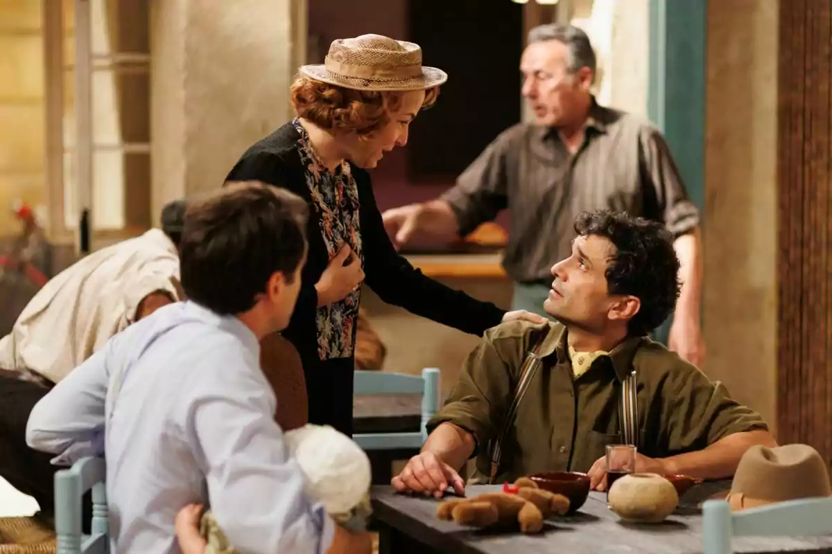 Teresa Hurtado d'Ory toca l'espatlla d'Andrea Fiorillo envoltat d'altres persones en una escena ambientada en un interior amb una taula i objectes quotidians a La Moderna. Teresa Hurtado d'Ory toca l'espatlla d'Andrea Fiorillo envoltat d'altres persones en una escena ambientada en un interior amb una taula i objectes quotidians a La Moderna.