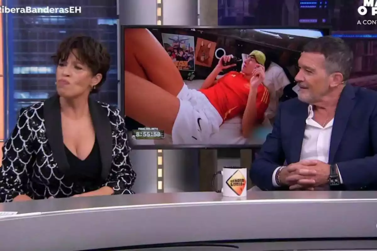Antonio Banderas y Marta Ribera están sentadas en el plató de El Hormiguero, con una pantalla detrás de ellos mostrando a una persona acostada con una camiseta roja y pantalones cortos blancos. Antonio Banderas y Marta Ribera están sentadas en el plató de El Hormiguero, con una pantalla detrás de ellos mostrando a una persona acostada con una camiseta roja y pantalones cortos blancos.