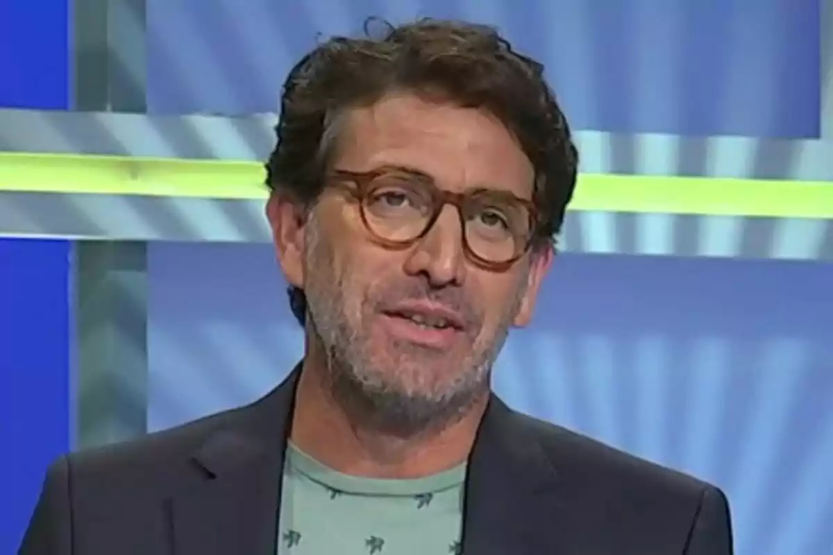 Antonio Garrido de cabello castaño y gafas marrones con barba y camiseta verde claro, de pie frente a un fondo azul con líneas amarillas y grises en Atrápame si puedes.