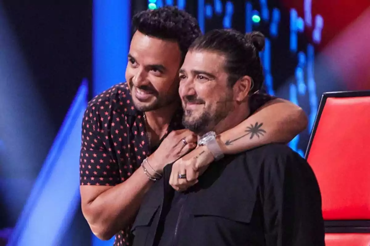 Antonio Orozco i Luis Fonsi somrients abraçant-se al plató de La Voz amb llums de fons. Antonio Orozco i Luis Fonsi somrients abraçant-se al plató de La Voz amb llums de fons.