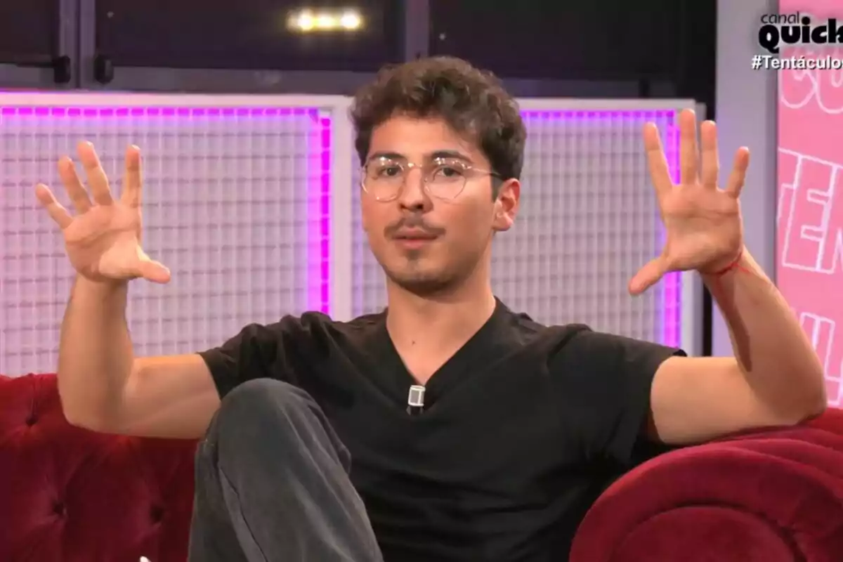 Arnau Martínez con gafas y camiseta negra está sentado en un sofá rojo con las manos levantadas en un gesto expresivo en Tentáculos iluminado con luces de neón. Arnau Martínez con gafas y camiseta negra está sentado en un sofá rojo con las manos levantadas en un gesto expresivo en Tentáculos iluminado con luces de neón.