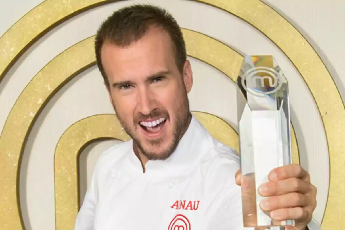 Arnau París con uniforme de chef sosteniendo un trofeo transparente frente a un fondo con un logotipo dorado en MasterChef. Arnau París con uniforme de chef sosteniendo un trofeo transparente frente a un fondo con un logotipo dorado en MasterChef.