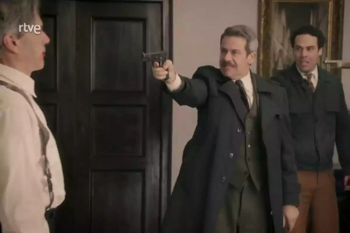 Juli Fábregas con bigote y traje oscuro apunta una pistola a Miguel Hermoso mientras Jesús Mosquera observa en una habitación con puertas de madera en La Moderna. Juli Fábregas con bigote y traje oscuro apunta una pistola a Miguel Hermoso mientras Jesús Mosquera observa en una habitación con puertas de madera en La Moderna.