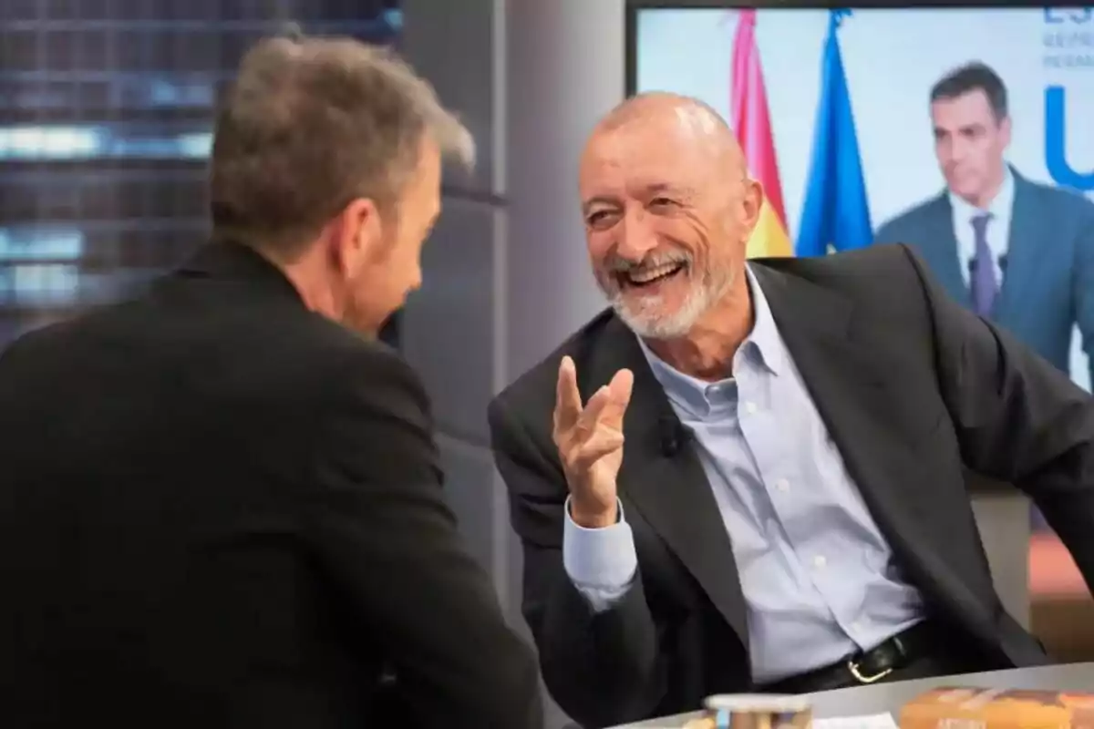 Arturo Pérez-Reverte y Pablo Motos conversando y sonriendo en el plató de El Hormiguero con una pantalla de fondo que muestra a Pedro Sánchez. Arturo Pérez-Reverte y Pablo Motos conversando y sonriendo en el plató de El Hormiguero con una pantalla de fondo que muestra a Pedro Sánchez.
