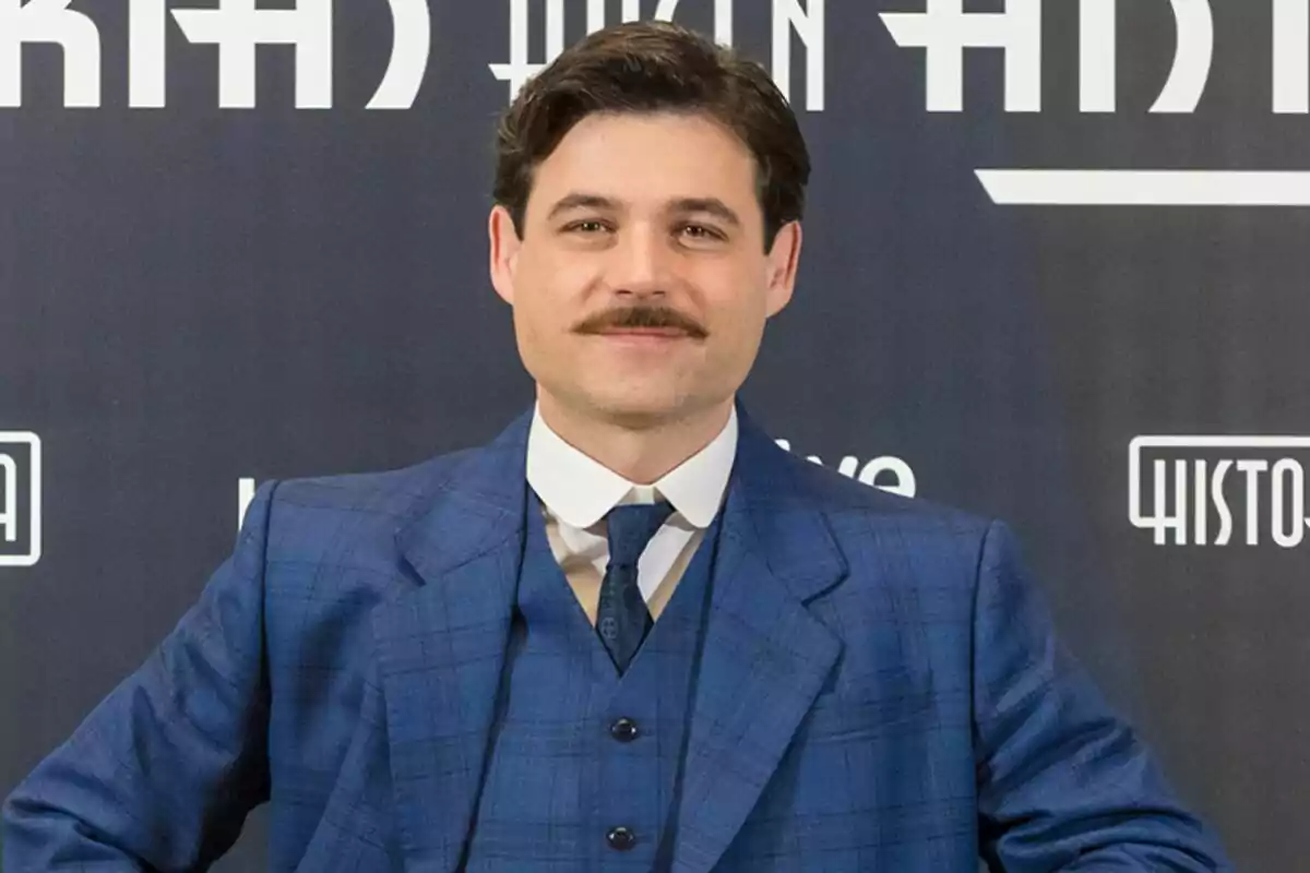 Arturo Sancho amb bigoti i vestit blau posant davant d'un fons amb text de La Promesa. Arturo Sancho amb bigoti i vestit blau posant davant d'un fons amb text de La Promesa.