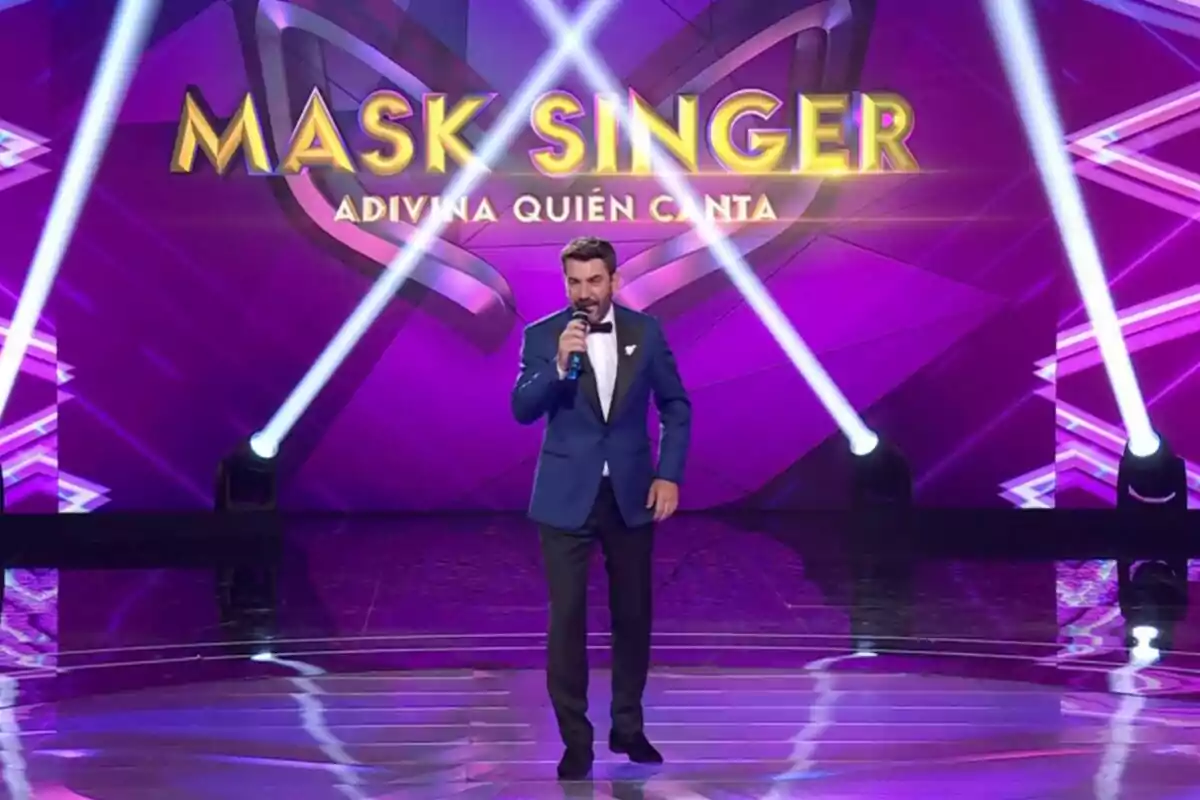 Un presentador amb vestit blau i corbatí negra sosté un micròfon a l'escenari del programa 