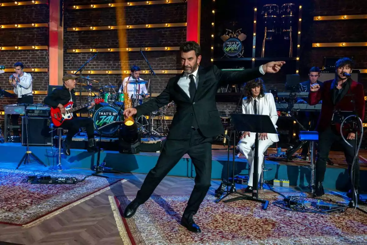 Arturo Valls amb vestit negre balla en un escenari mentre una banda toca música al fons, amb llums brillants i una atmosfera animada a That's my jam.