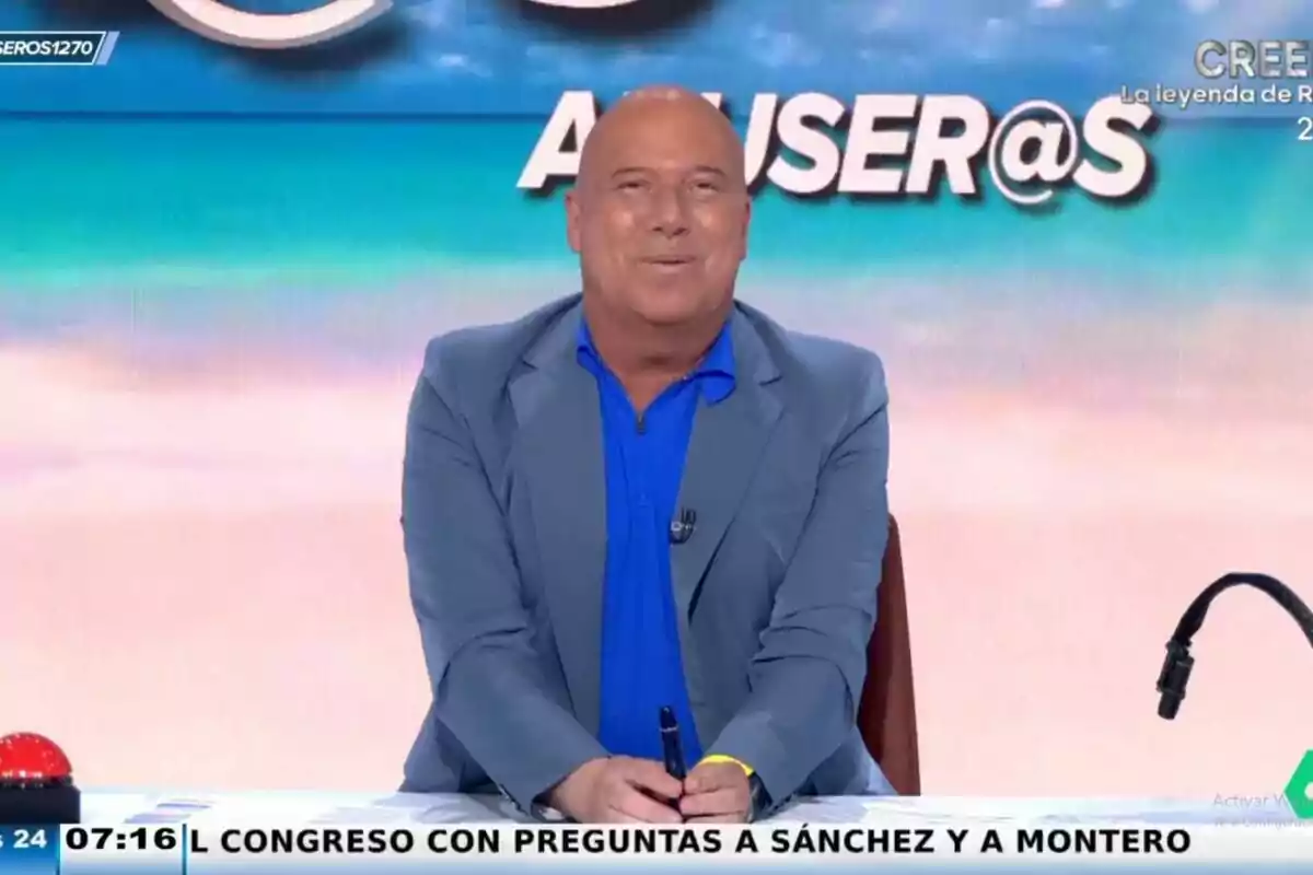 Alfonso Arús con una chaqueta azul claro y camisa azul está sentado en el set de 'Aruser@s' con un fondo colorido y un letrero que dice Alfonso Arús con una chaqueta azul claro y camisa azul está sentado en el set de 'Aruser@s' con un fondo colorido y un letrero que dice