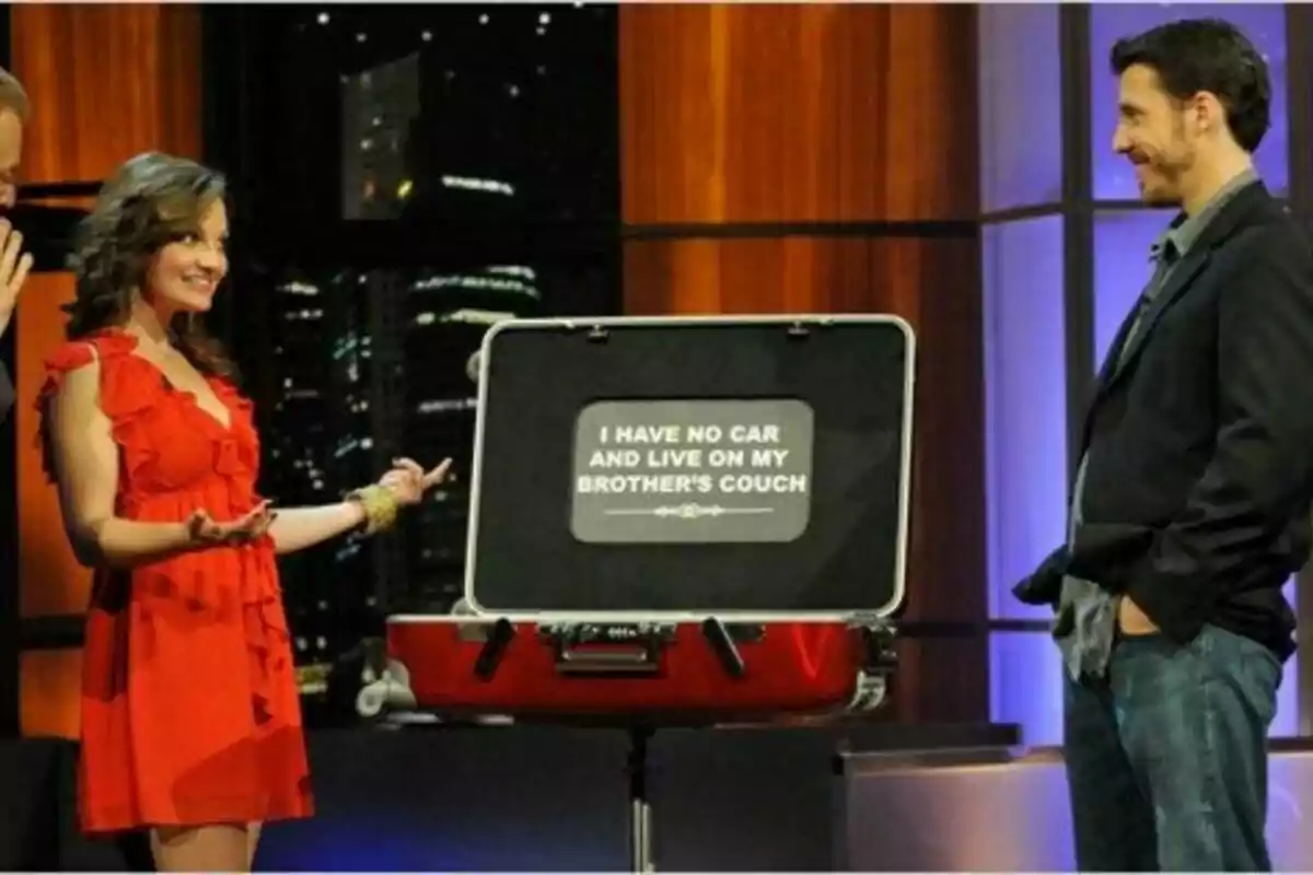 Una mujer con vestido rojo y un hombre con chaqueta se miran sonrientes en un set de televisión de Baggage junto a una maleta abierta que muestra un letrero en inglés que dice que no tiene auto y vive en el sofá de su hermano Una mujer con vestido rojo y un hombre con chaqueta se miran sonrientes en un set de televisión de Baggage junto a una maleta abierta que muestra un letrero en inglés que dice que no tiene auto y vive en el sofá de su hermano