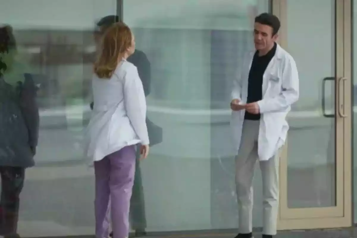Bahar y Timur con batas blancas conversan frente a un edificio con puertas de vidrio en Renacer.