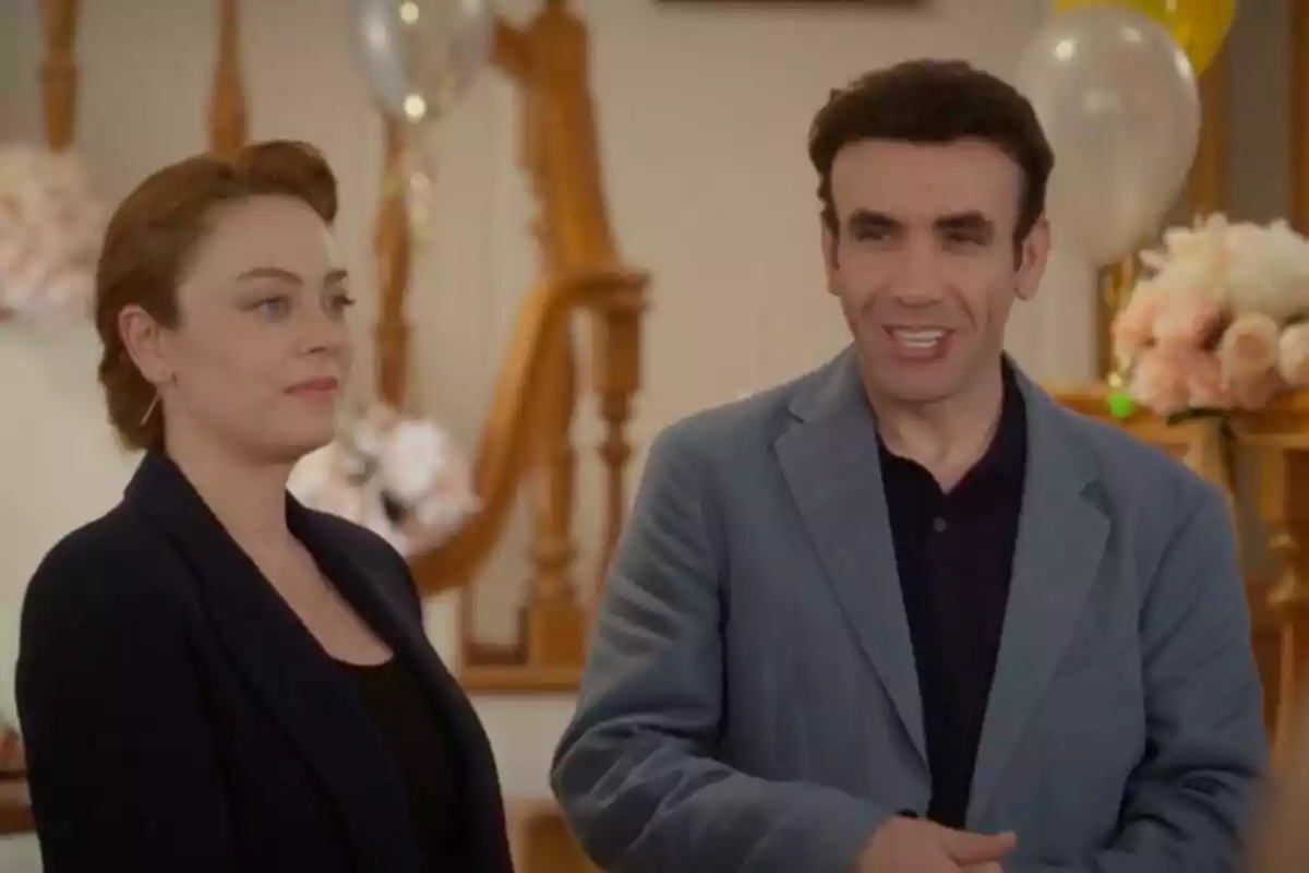 Bahar y Timur de pie en un ambiente decorado con globos y flores, una mujer con cabello recogido y un hombre con chaqueta azul.