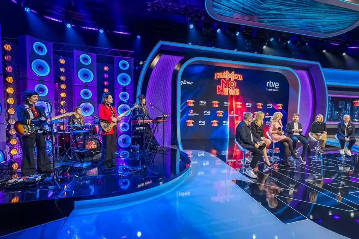 Un grupo musical toca en un escenario iluminado, 'José Mota No News' con luces azules y púrpuras mientras varias personas están sentadas en sillas en un set de televisión. Un grupo musical toca en un escenario iluminado, 'José Mota No News' con luces azules y púrpuras mientras varias personas están sentadas en sillas en un set de televisión.