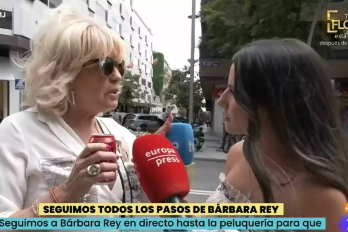 Dos mujeres conversan en la calle mientras Bárbara Rey sostiene una lata y varias reporteras les acercan micrófonos de diferentes medios de comunicación como La Familia de la Tele